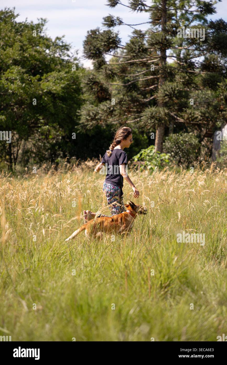 Une jeune femme s'amuse à jouer avec son chien dans un champ rempli d'herbes hautes. Ils courent ensemble, profitant du plein air sur une journée ensoleillée lumineuse environnante Banque D'Images