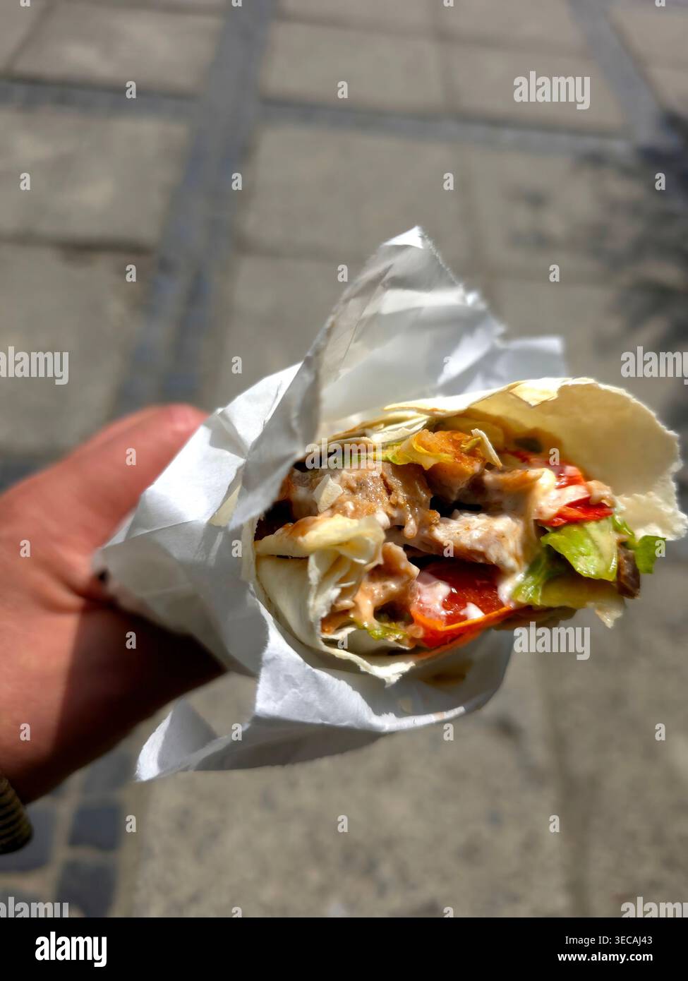 Une personne tient un kebab enveloppé dans sa main tout en se tenant debout sur le trottoir. Le kebab comprend de la viande, de la laitue et de la sauce. Banque D'Images