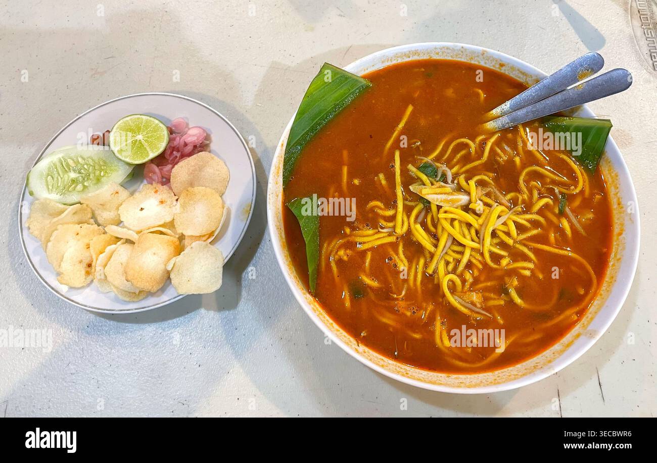 Un bol vibrant de Mie Aceh indonésien, riche en bouillon épicé et nouilles jaunes, servi avec du concombre frais, du citron vert, des échalotes marinées et du vidage Banque D'Images