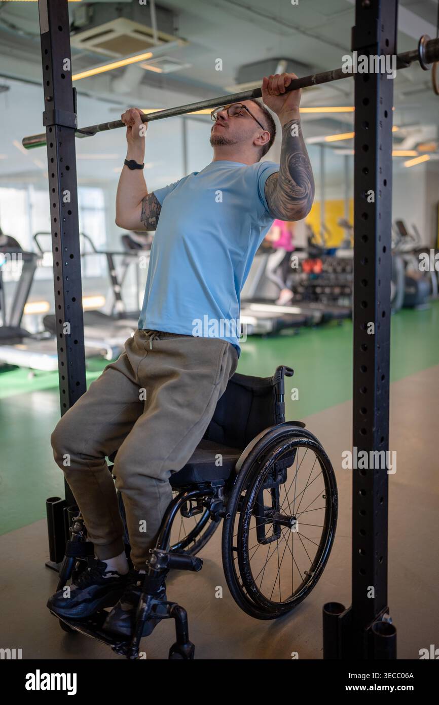Homme athlète en fauteuil roulant faisant des exercices verticaux de traction en utilisant un équipement sportif adaptatif au gymnase. Banque D'Images