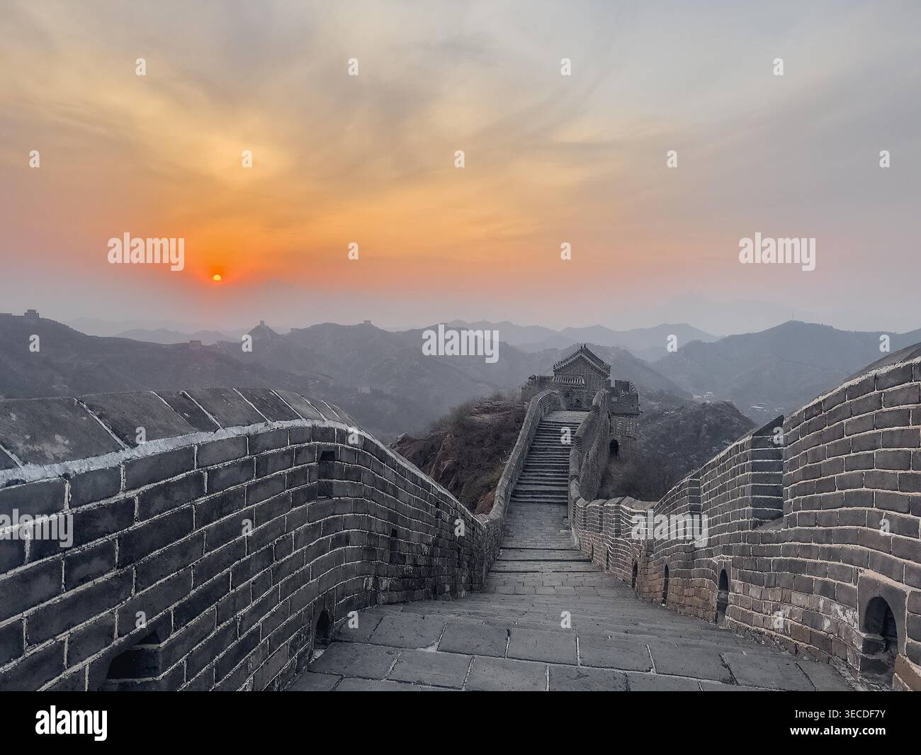 Le coucher du soleil brille sur d'anciens murs de pierre. Des escaliers mènent vers le haut à travers des pics montagneux accidentés. La lumière dorée peint le ciel dans des teintes ambre douces. Des tours lointaines se dressent fièrement au milieu de collines brumeuses Banque D'Images
