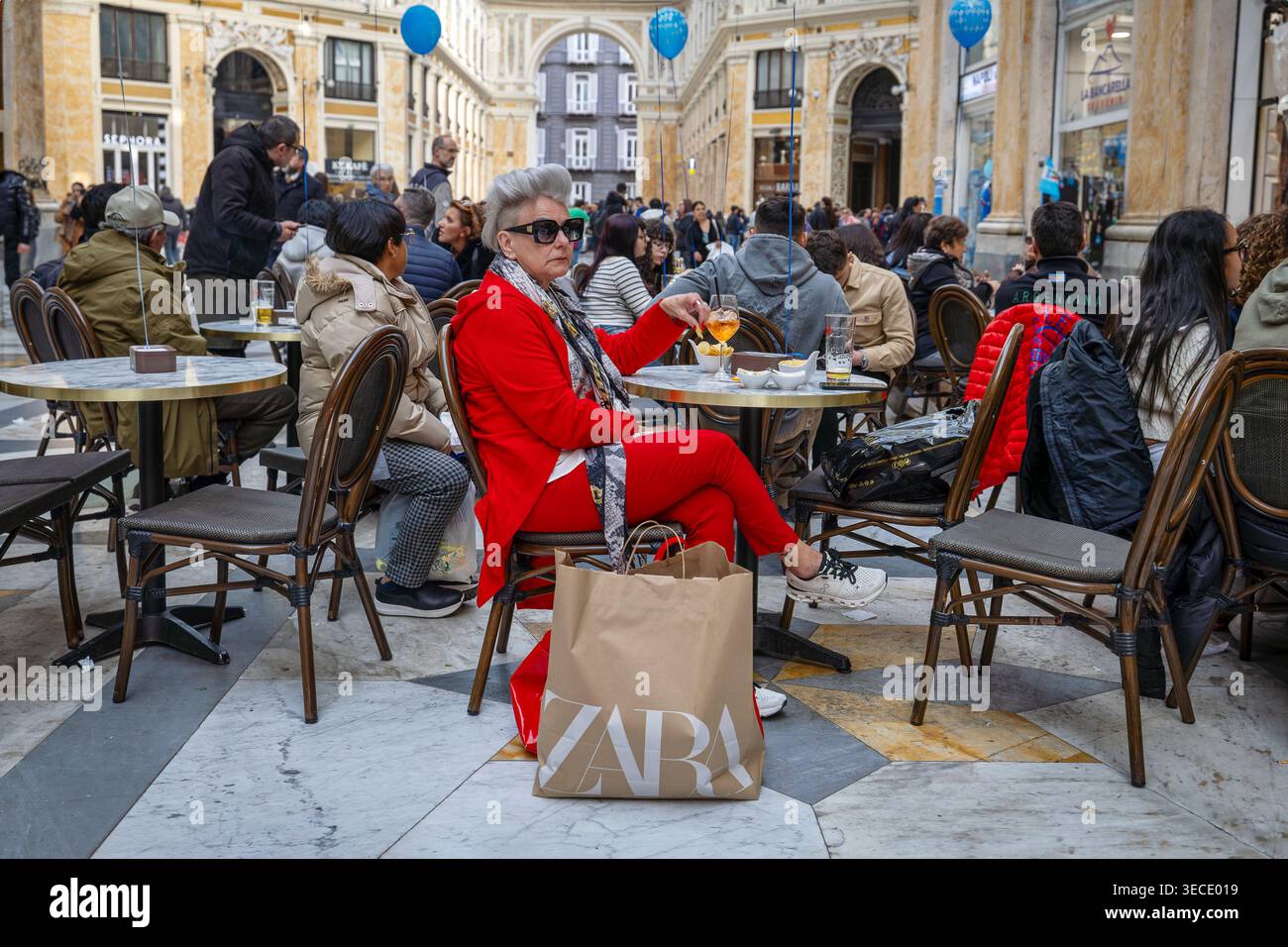 Naples, Italie - 19 mars 2026 : une femme élégante avec des cheveux argentés et un costume rouge vif prend l'apéritif à une table de café dans l'historique Galleria UMB Banque D'Images