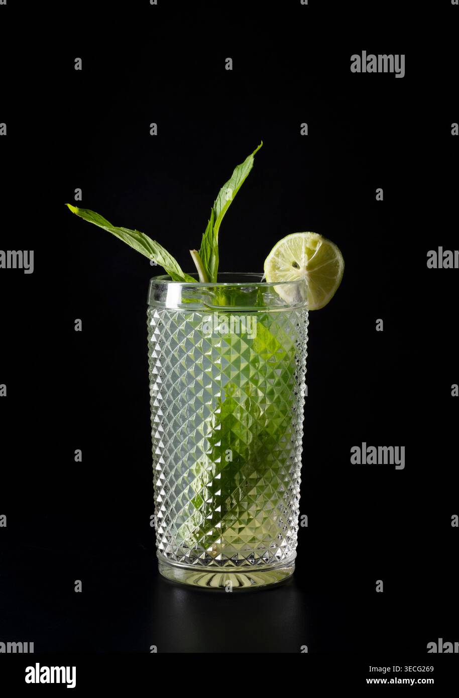 Cocktail mojito rafraîchissant servi dans un verre texturé avec des feuilles de menthe fraîche et une tranche de citron vert sur fond noir. Photographie de studio minimale Banque D'Images