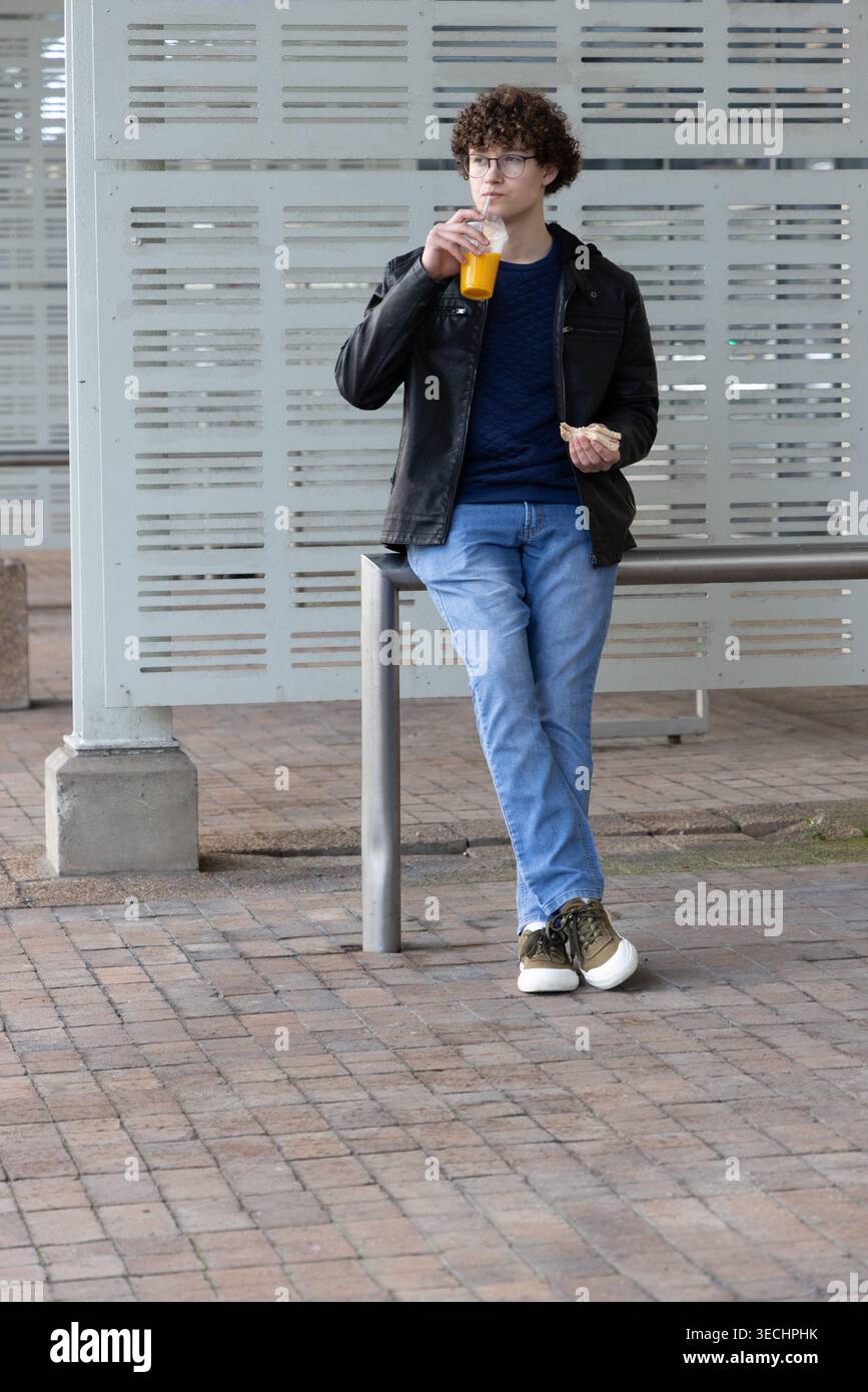 Homme appuyé sur un rail métallique dans la salle d'attente abritée, tenant une boisson à l'orange et de la pâtisserie Banque D'Images