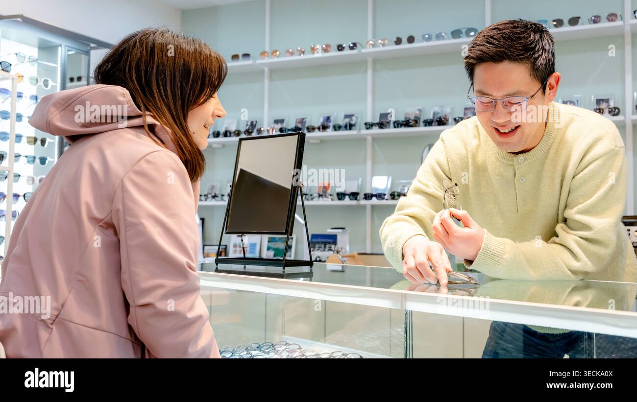Un opticien amical assiste un client dans un centre optique, en présentant diverses lunettes. L'interaction met l'accent sur la satisfaction et le perso du client Banque D'Images
