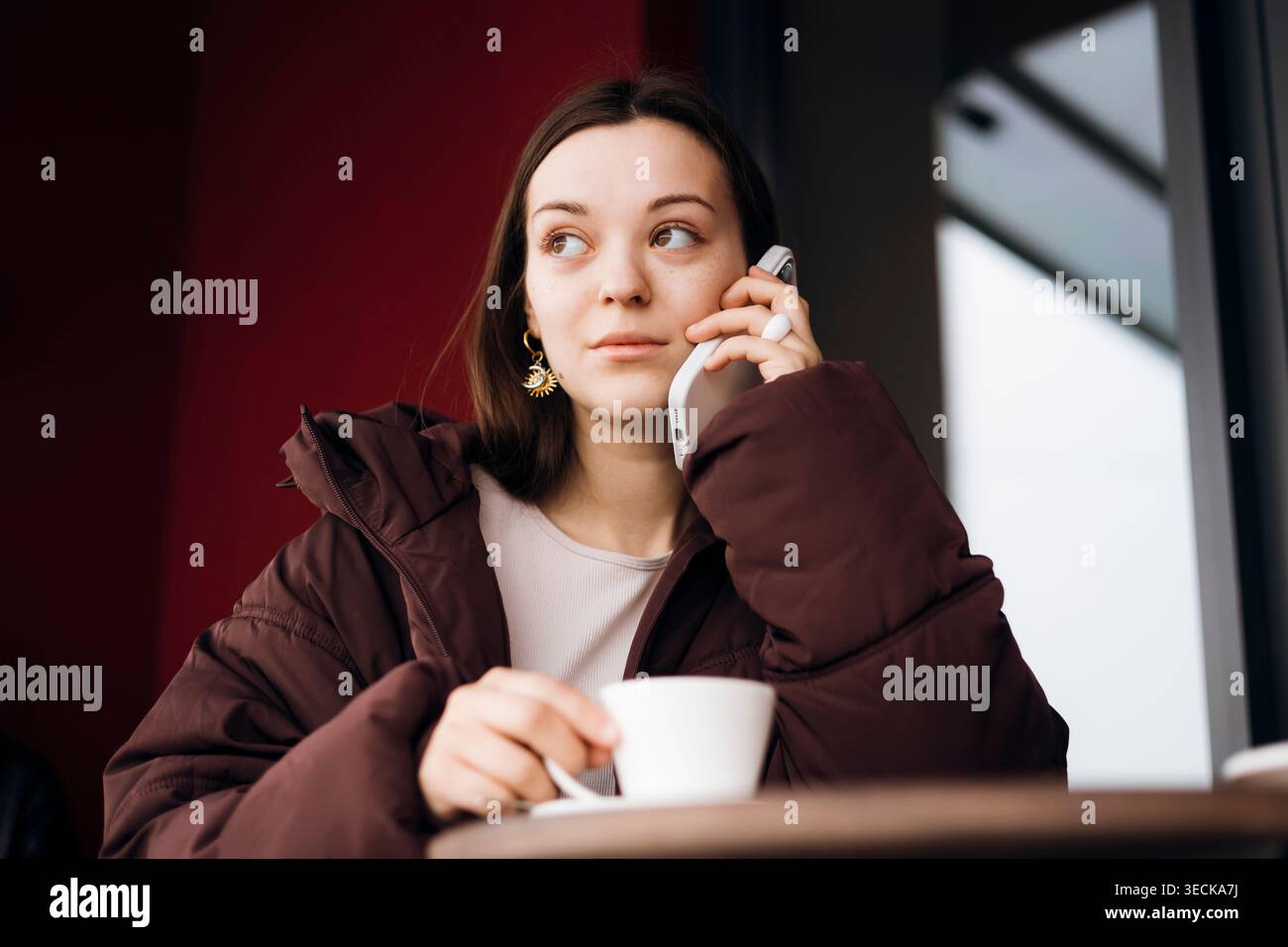 Jeune femme réfléchie dans une veste brune parle sur un smartphone tout en tenant une tasse de café à une table de café, regardant de côté près d'une fenêtre avec da doux Banque D'Images