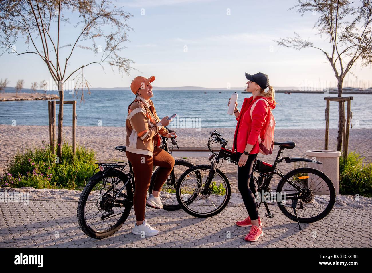 Deux amies sportives font une pause sur une promenade en bord de mer avec des vélos électriques, bavardant et s'hydratant après une balade, profitant du temps ensoleillé et en plein air Banque D'Images