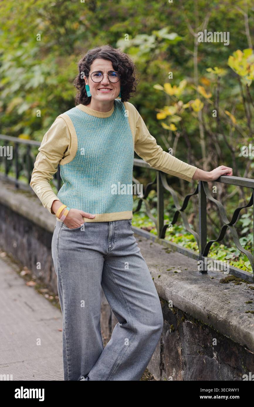Femme mature avec des cheveux bouclés et des lunettes, vêtue de façon décontractée, profite d'un moment de détente appuyé sur une balustrade de fer dans un cadre verdoyant de parc urbain, EM Banque D'Images