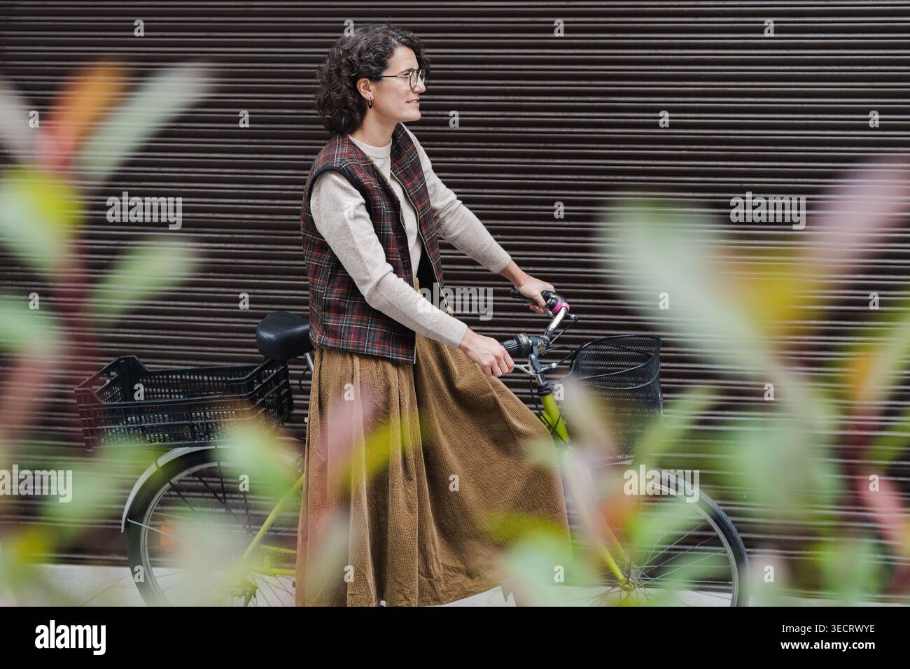 Femme mature avec les cheveux bouclés et les lunettes marche le long de sa bicyclette dans un cadre urbain. Elle est habillée dans des tons automnaux et un gilet à carreaux, appréciant Banque D'Images
