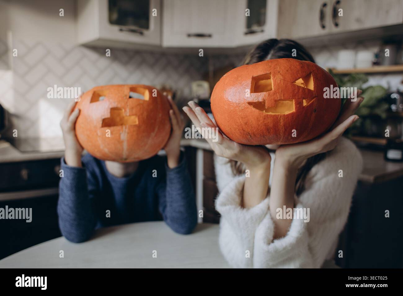 Frère et sœur jouent à la maison, cachant leurs visages derrière des lanternes de citrouille sculptées, créant une ambiance amusante pour Halloween avec une atmosphère d'automne intérieure confortable Banque D'Images