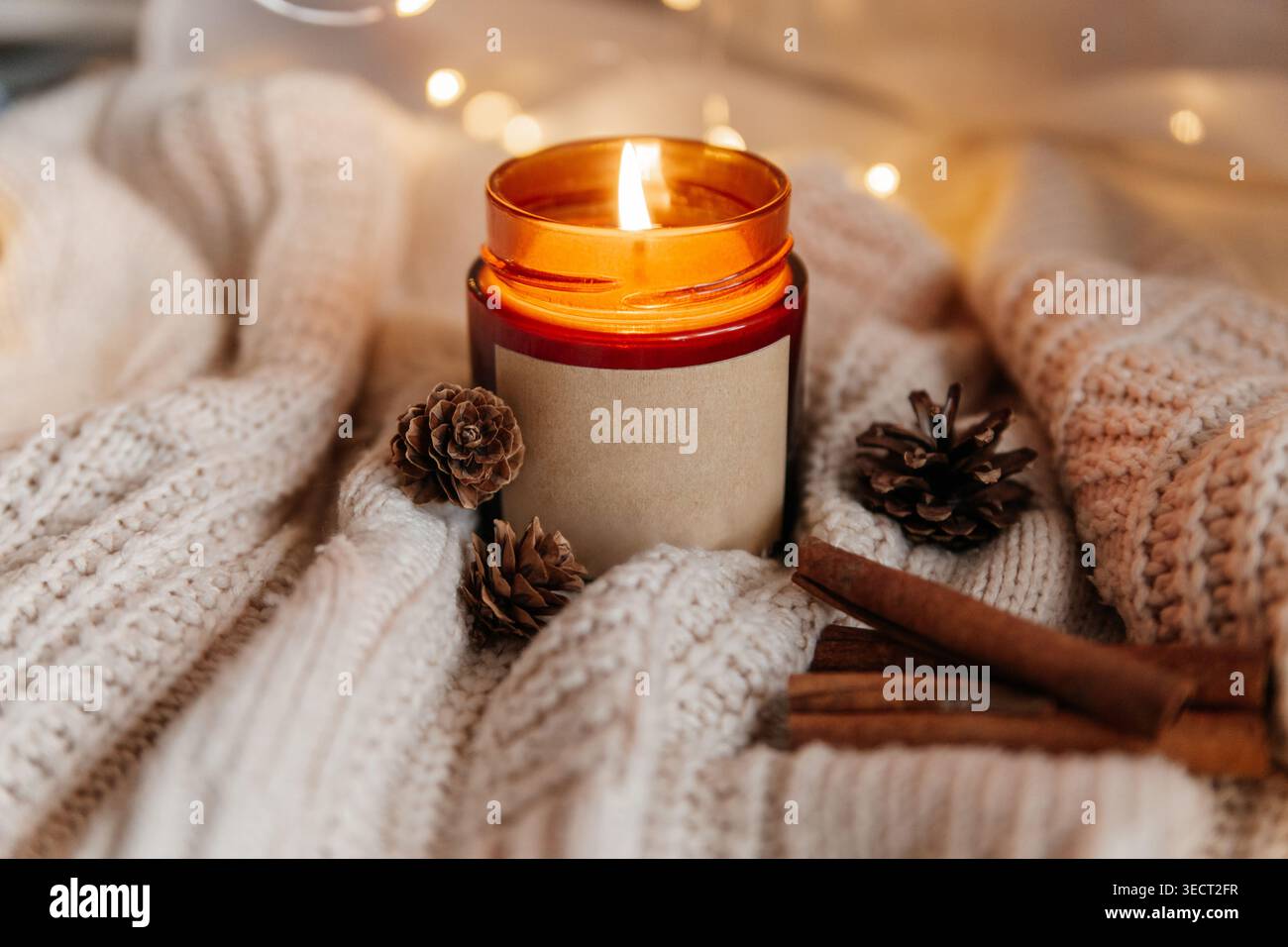 Bougie en pot ambre chaude brûlant sur une couverture tricotée douce, stylisée avec des pommes de pin et des bâtons de cannelle. Hygge d'hiver confortable nature morte avec ligh de fée bokeh Banque D'Images