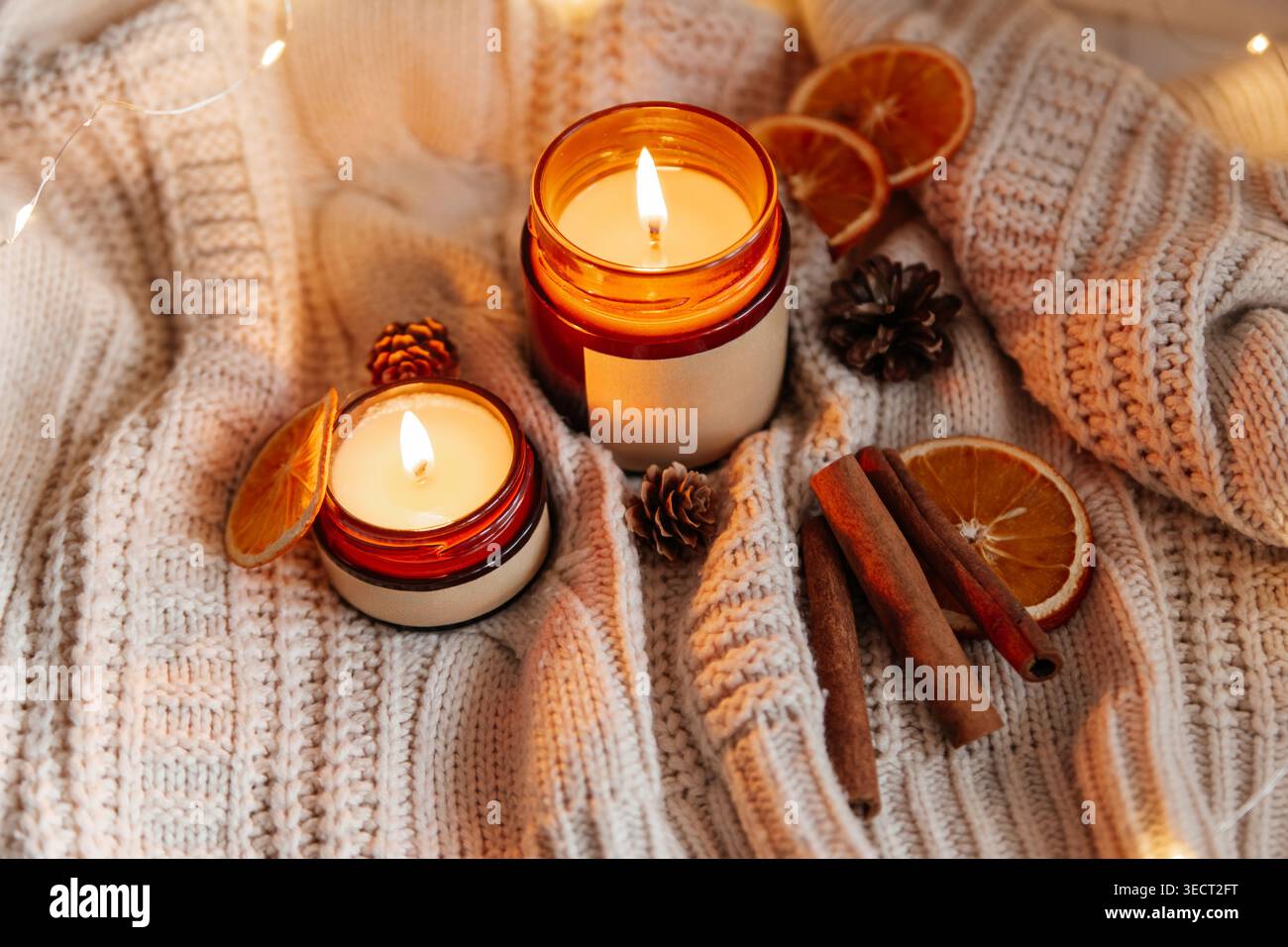 Deux bougies de soja incandescentes dans des pots ambrés sur une couverture en grosse maille, stylisées avec des tranches d'orange séchées, des bâtonnets de cannelle et des pommes de pin pour un hygge hivernal chaud Banque D'Images