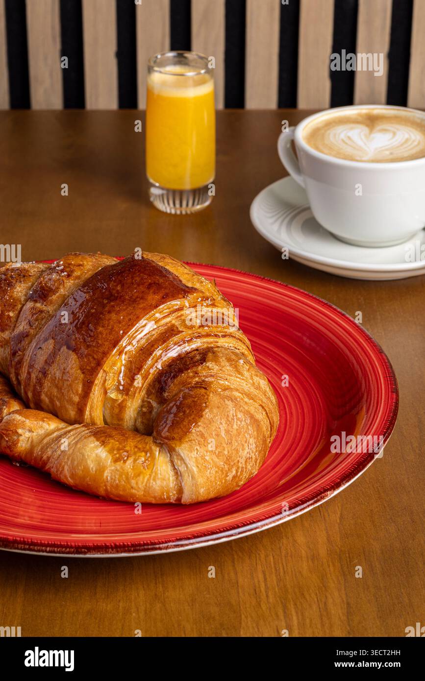 Petit déjeuner frais sur une table en bois avec un croissant feuilleté sur une assiette rouge, latte crémeux avec art latte coeur, et un verre de jus d'orange. Banque D'Images