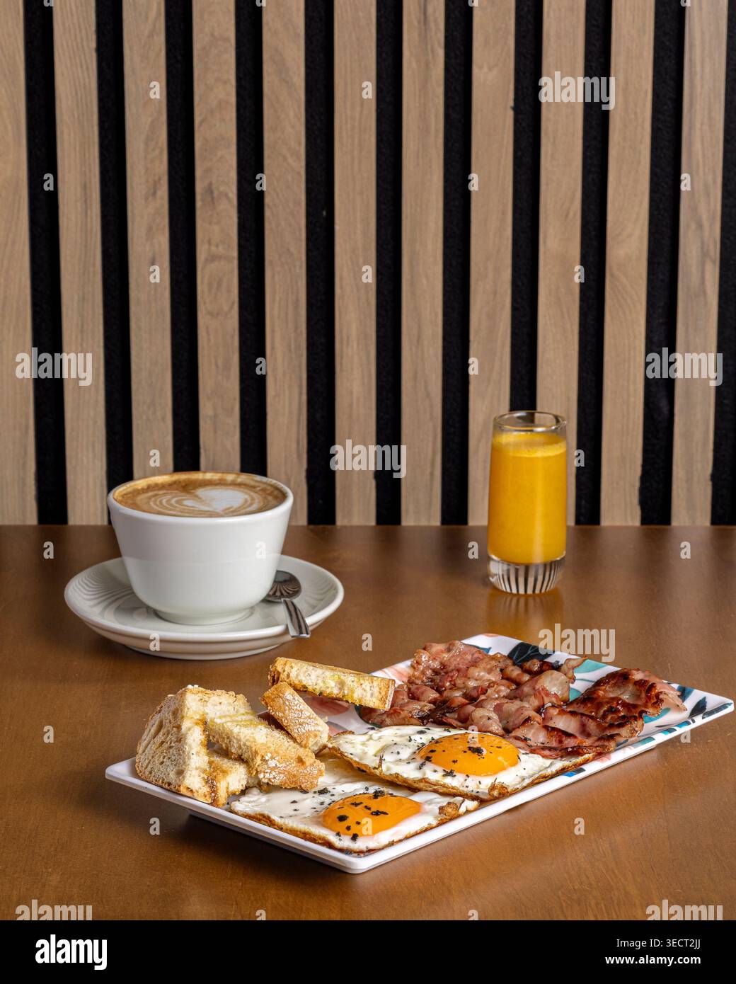 Petit-déjeuner matinal sur une table avec un oeuf frit avec pain grillé, bacon croustillant et jambon, un cappuccino dans une tasse blanche, et un verre de jus d'orange dans un coz Banque D'Images