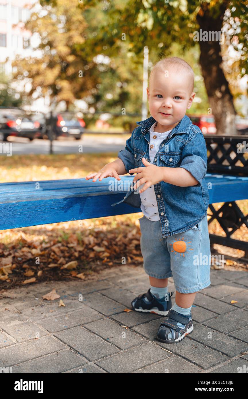 Mignon tout-petit dans une veste en Jean et un short se tient près d'un banc bleu dans un parc d'automne ensoleillé, souriant à la caméra avec des arbres flous et la rue dans la BA Banque D'Images
