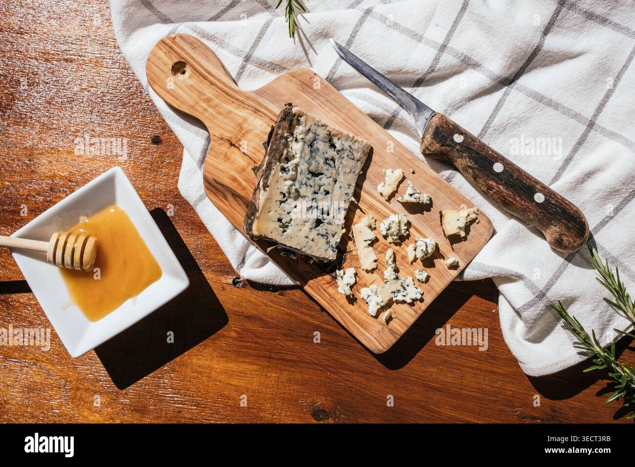 Scène culinaire rustique avec fromage bleu sur une planche à découper en bois, morceaux émiettés, un couteau vintage, brins de romarin et un bol de miel sur un tex Banque D'Images