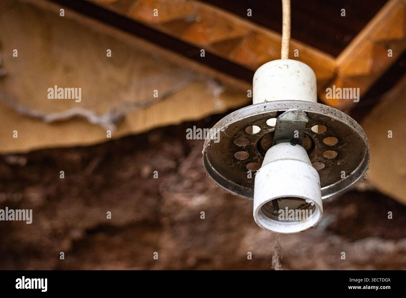 Vue rapprochée d'une vieille suspension avec douille en céramique poussiéreuse et abat-jour en métal perforé suspendu à un plafond fatigué. L'âge et les toiles d'araignée suggèrent un aband Banque D'Images