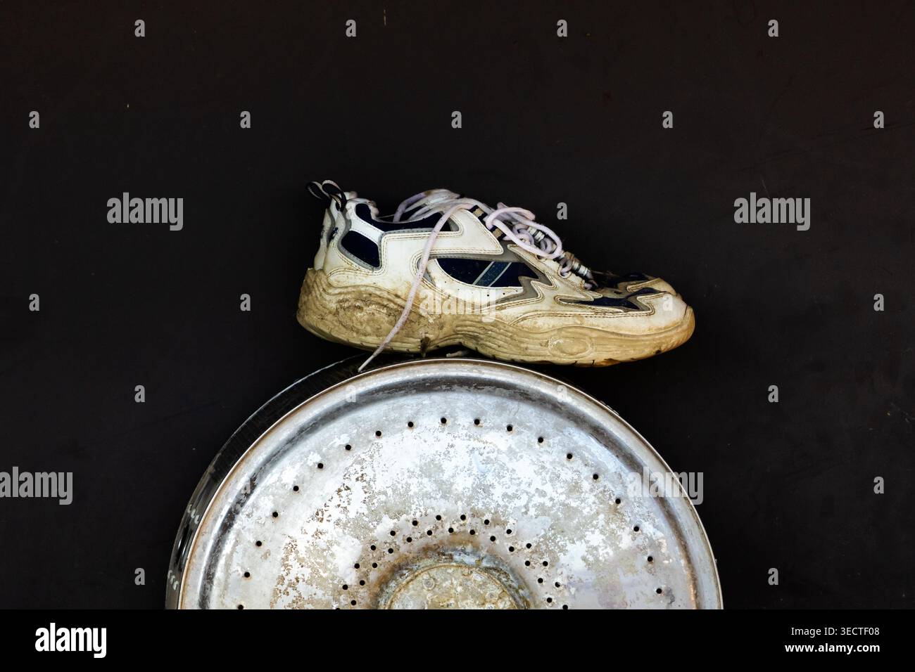 La vieille chaussure de sport repose sur une plaque métallique altérée sur un fond sombre mettant en évidence les taches de saleté sur les semelles éraflées et le charme des objets trouvés urbains dans un qu Banque D'Images