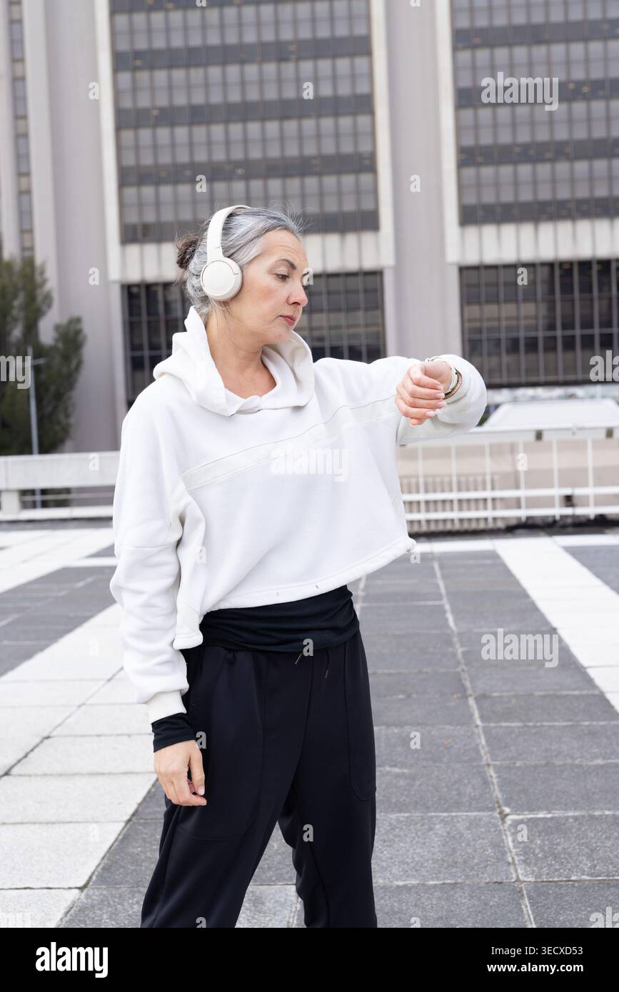 Femme mature debout sur la terrasse sur le toit vérifiant le dispositif de poignet, portant des écouteurs et un sweat à capuche blanc Banque D'Images