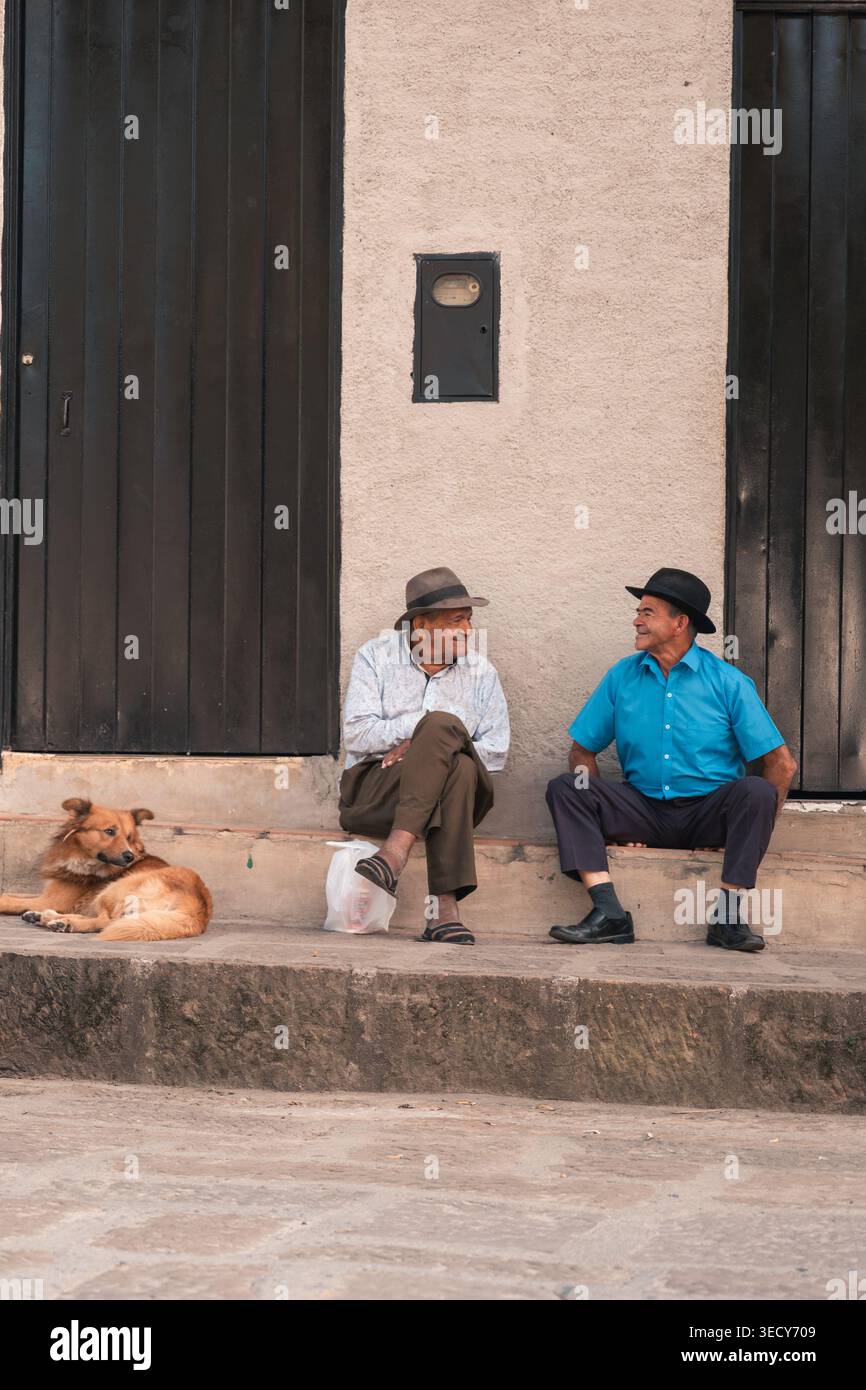 Mogotes, colombie - marzo 21, 2026 deux hommes plus âgés sont assis sur les marches d'une petite ville colombienne, bavardant et souriant tandis qu'un chien se repose à côté d'eux, rel Banque D'Images