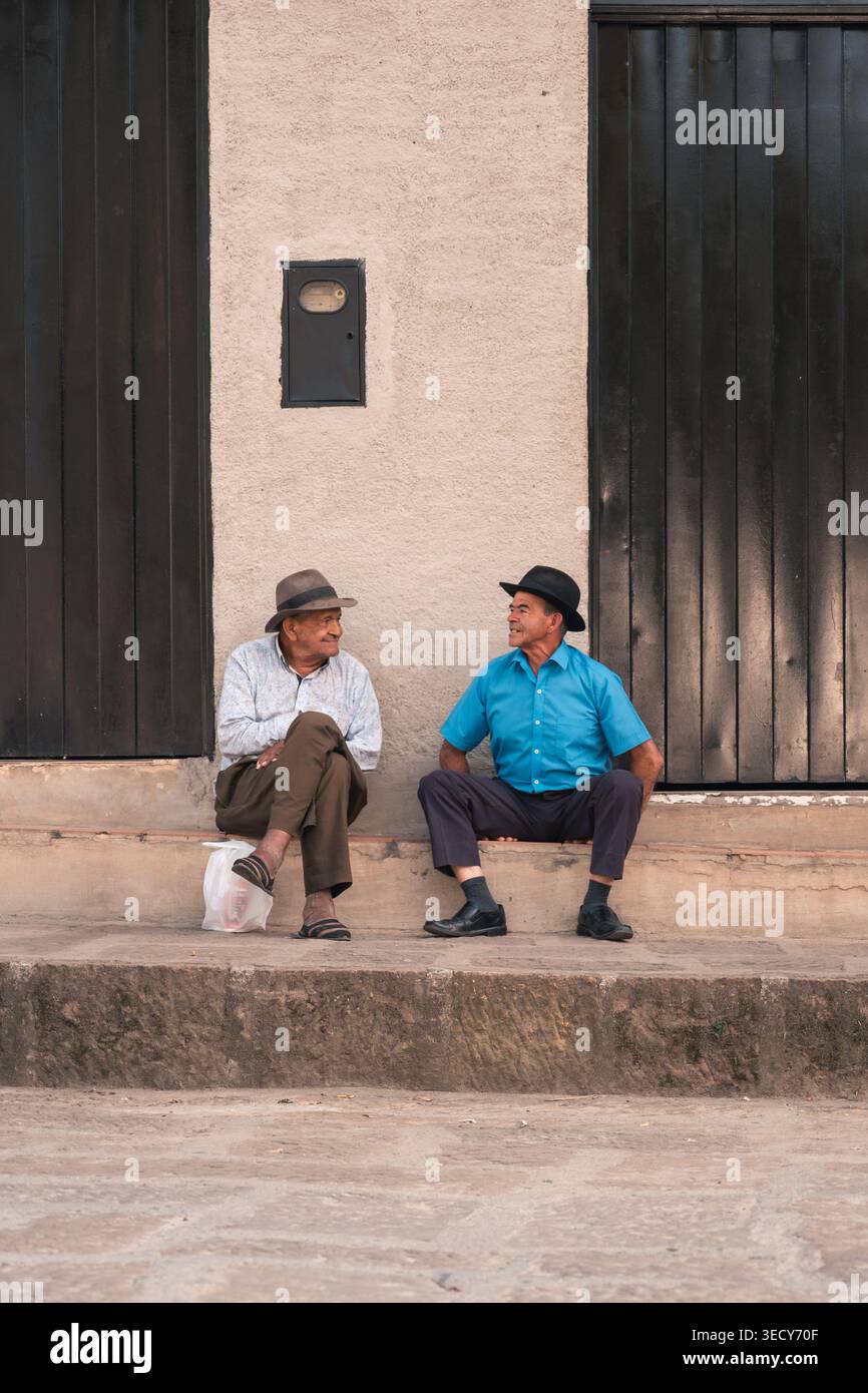 Mogotes, colombie - marzo 21, 2026 deux hommes hispaniques âgés en chapeaux sont assis sur un trottoir, discutant et regardant la vie de la rue dans un colome urbain détendu Banque D'Images