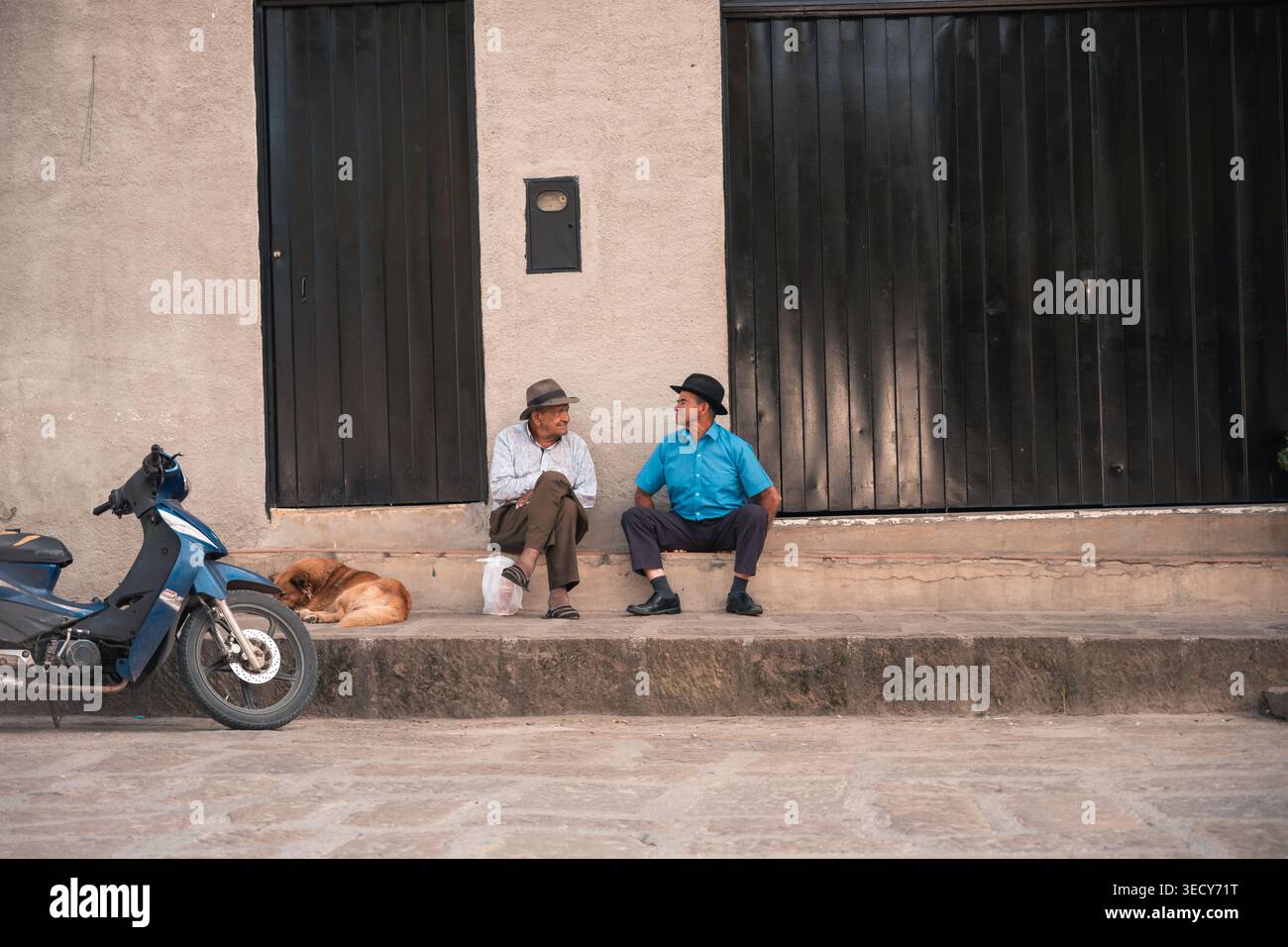 Mogotes, colombie - marzo 21, 2026 deux adultes âgés assis sur des marches, discutant à l'extérieur d'un bâtiment avec une moto bleue et un chien endormi Banque D'Images