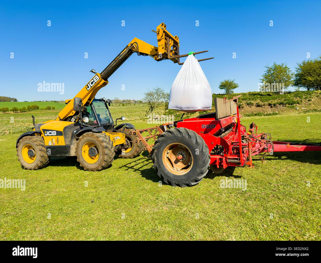 Chargeur à bras télescopique JCB jaune chargeant un grand sac blanc de semences en vrac dans une planteuse agricole rouge dans un champ vert sous un ciel bleu clair Banque D'Images