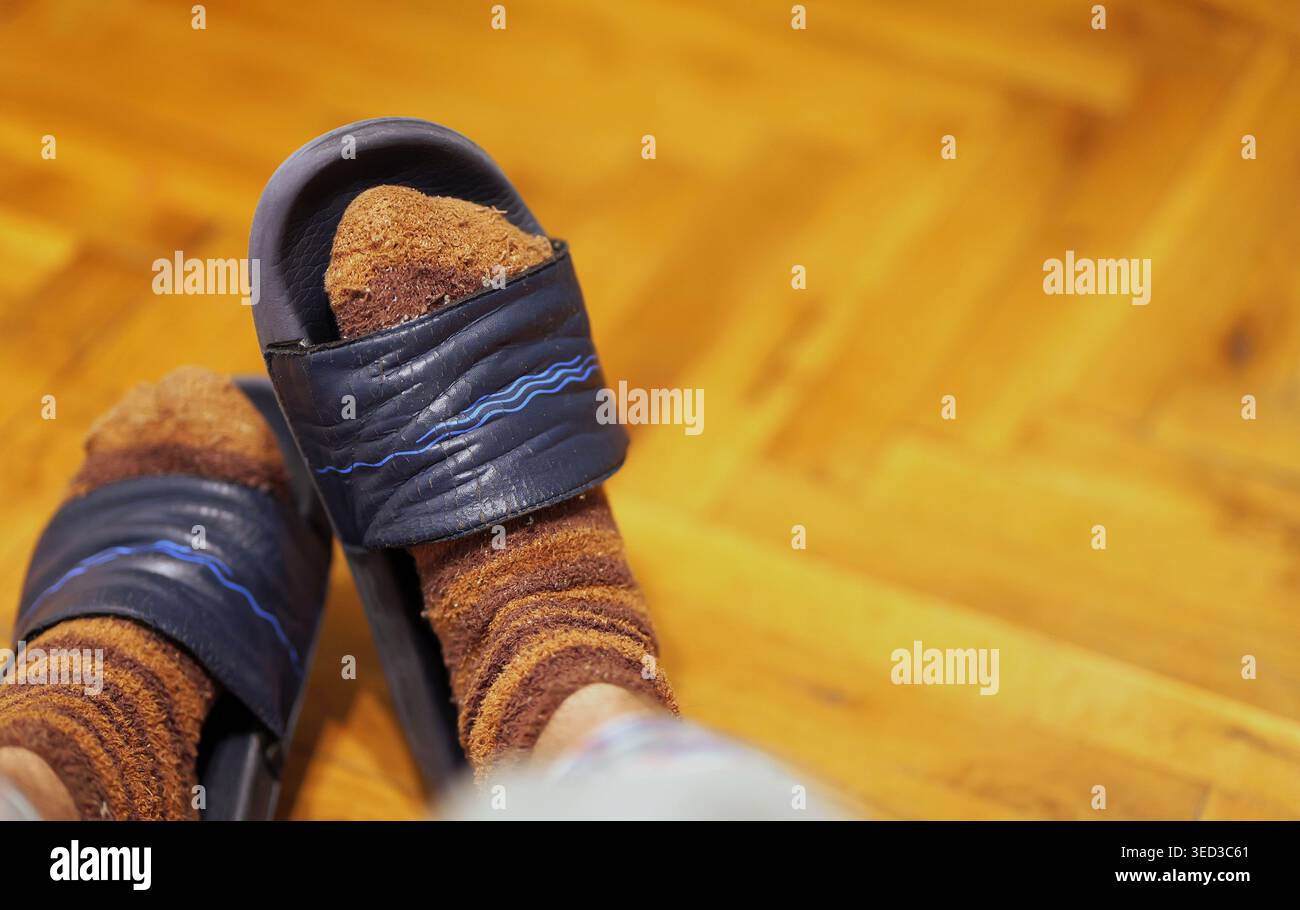 Gros plan de pieds portant des chaussettes tricotées marron et des pantoufles coulissantes bleu foncé sur un parquet en bois, créant une atmosphère chaleureuse et décontractée à la maison. Banque D'Images