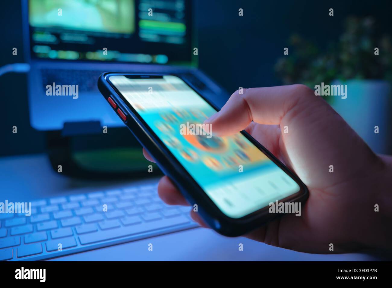 Gros plan de la main tenant un smartphone et faisant défiler rapidement le flux de médias sociaux dans une pièce sombre avec un bureau sur fond, éclairé par une lumière bleue. Dépendance numérique et consommation de contenu sans esprit Banque D'Images