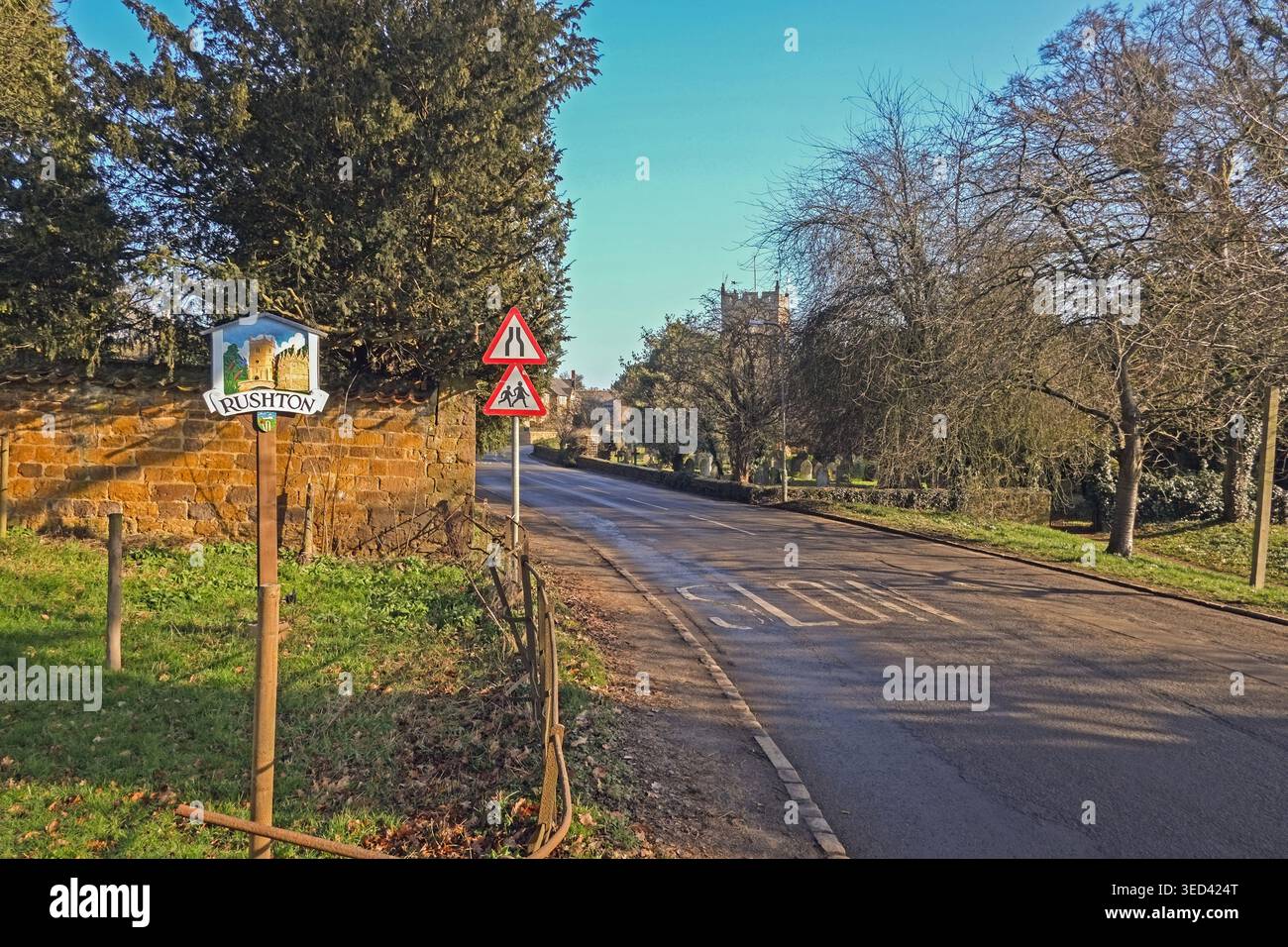 Route menant au village de Rushton dans le Northamptonshire avec un panneau de rue, des arbres et une église en arrière-plan pendant les heures de jour Banque D'Images