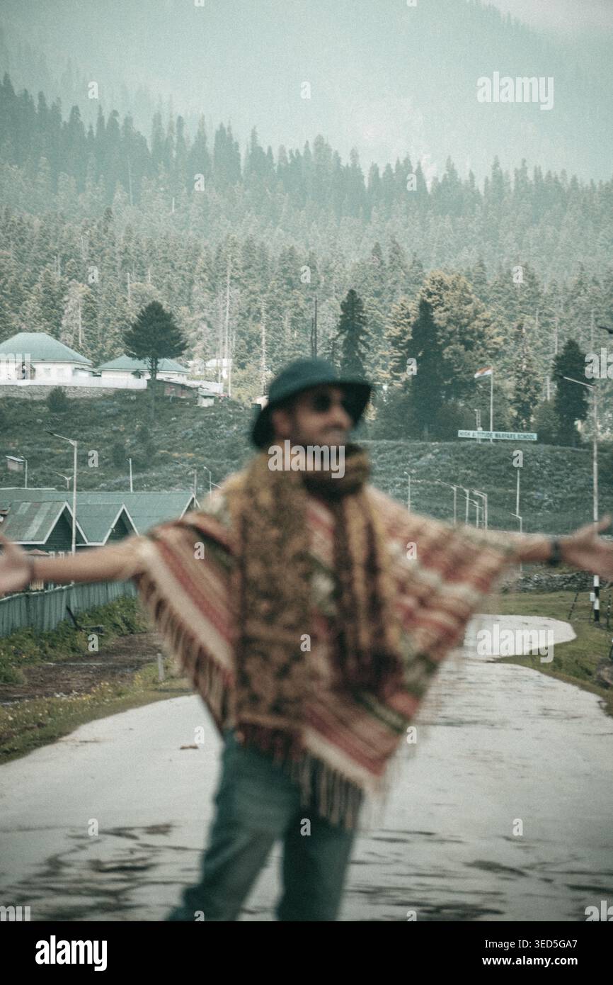 Un homme portant un poncho à motifs et un chapeau se tient debout avec ses bras tendus sur une route humide dans un paysage de montagne pittoresque avec des pins et un bâtiment Banque D'Images