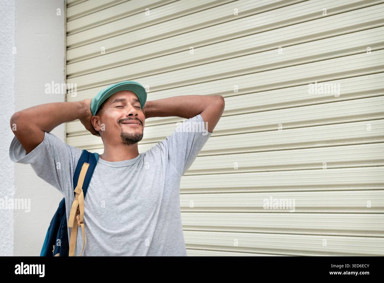 Homme souriant avec les yeux fermés et les mains derrière la tête, se sentant satisfait en milieu urbain Banque D'Images