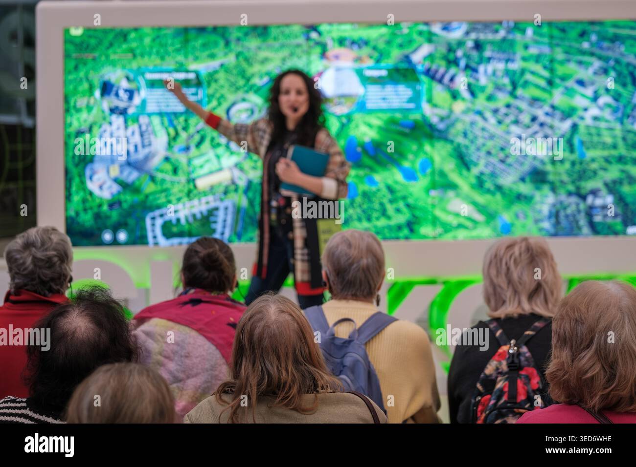 Une conférencière féminine présente une carte de planification urbaine au public assis à l'exposition, les aînés écoutent attentivement, la visite éducative, l'affichage de la technologie, l'engagement communautaire. Banque D'Images