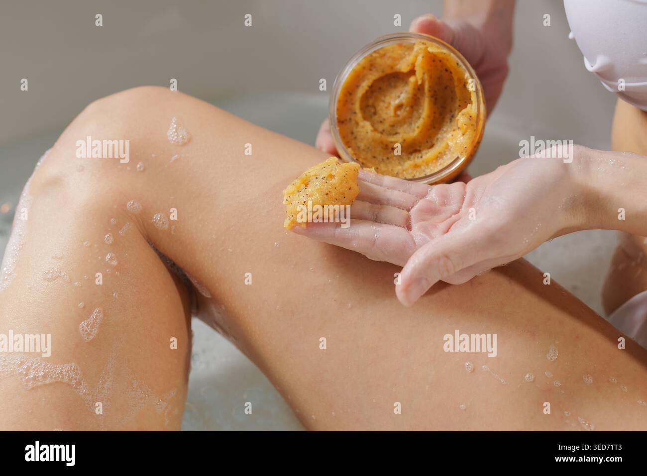 Une femme applique un gommage corporel naturel à sa jambe tout en profitant d'un bain relaxant, mettant en valeur les routines d'auto-soin et de soin de la peau pour une peau saine. Banque D'Images