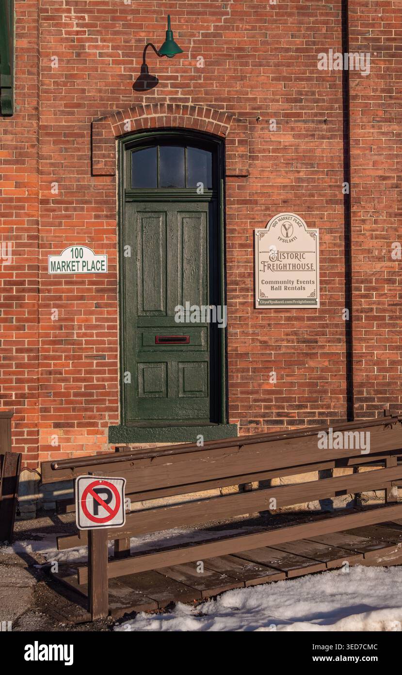 Ypsilanti, États-Unis - 12 février 2026 : façade historique de bâtiment en briques rouges avec porte voûtée verte, lampes murales vintage, panneaux de signalisation et neige, petite ville wi Banque D'Images