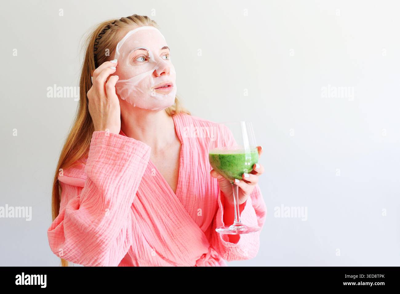 Femme en peignoir rose appliquant le masque de feuille et tenant le smoothie vert, routine beauté spa maison. Banque D'Images
