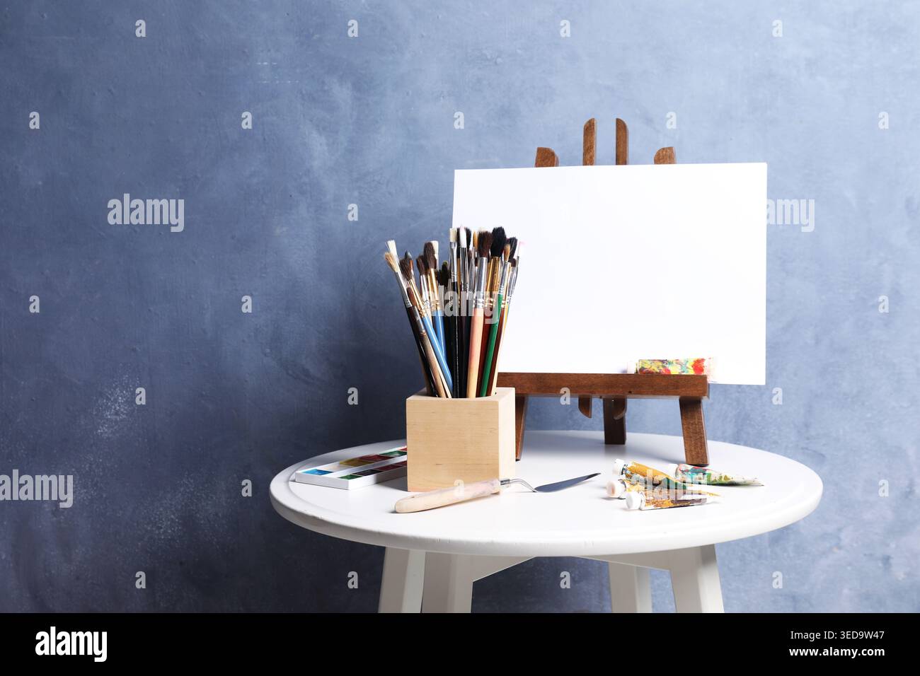 Pinceaux, couteau, peintures et chevalet avec toile sur table près du mur bleu. Espace pour le texte Banque D'Images