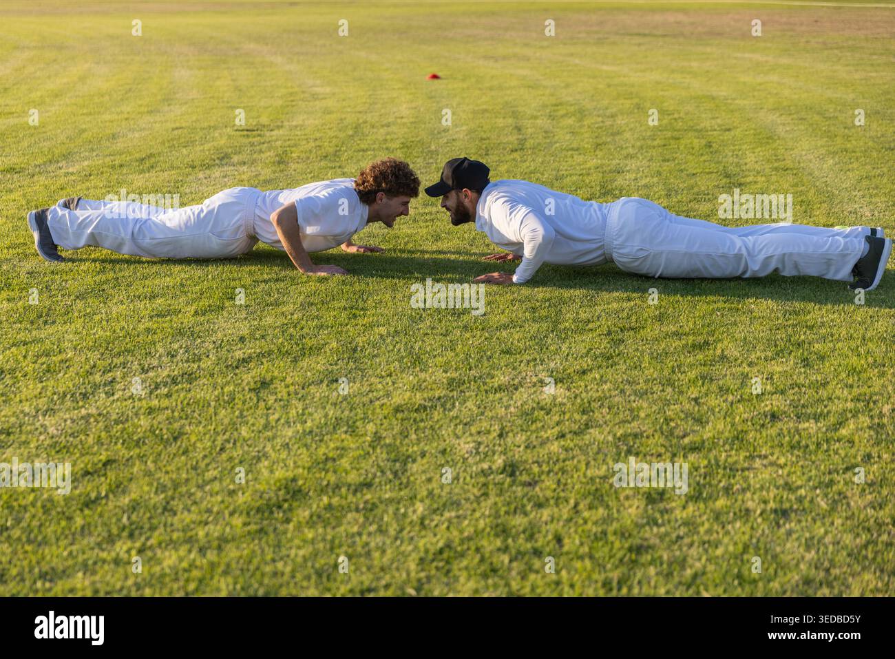 Deux hommes effectuant des pompes sur terrain en herbe en uniformes blancs avec cône d'entraînement orange à proximité Banque D'Images