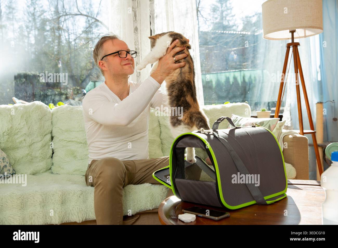 Homme mûr plaçant le chat dans le transporteur à la maison, propriétaire responsable de l'animal de compagnie, visite vétérinaire, voyage de l'animal de compagnie et routine de soins quotidiens dans le salon lumineux. Banque D'Images