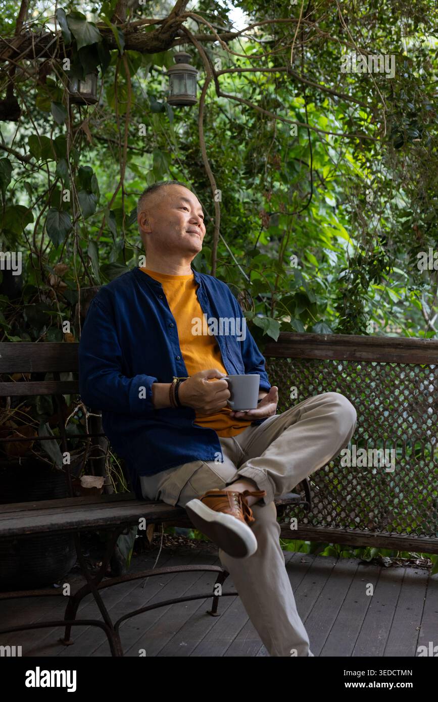 Homme asiatique senior assis sur un banc en bois sur un porche couvert tenant une tasse en céramique, portant une chemise marine Banque D'Images