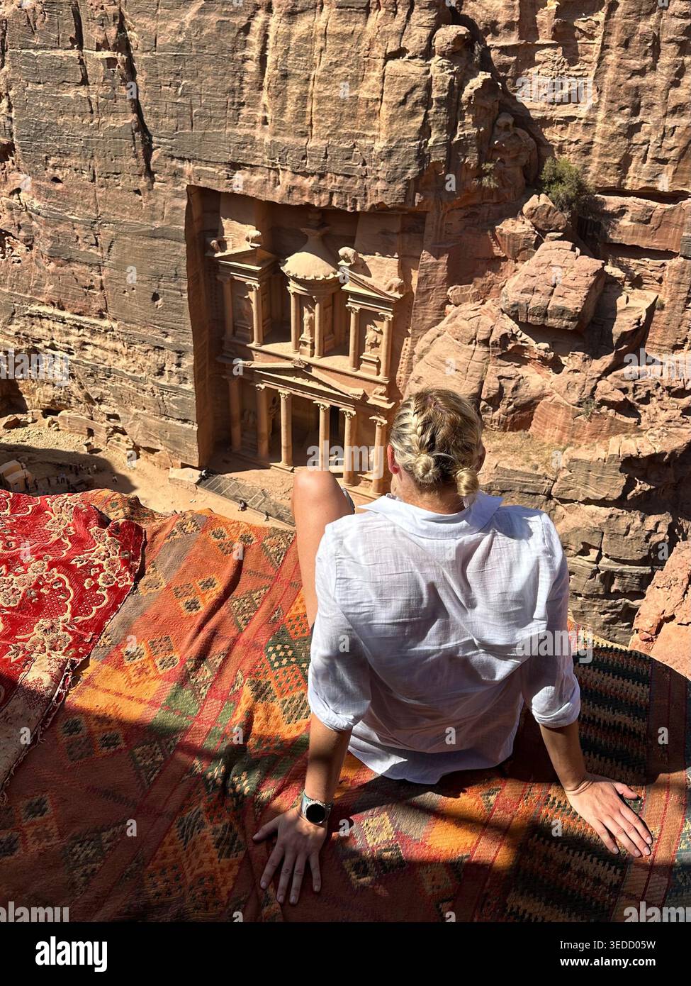 Une personne assise sur une corniche rocheuse surplombant le Trésor à Petra, en Jordanie, avec une vue panoramique sur l'ancienne ville sculptée ci-dessous. - Image de stock capturée avec un smartphone