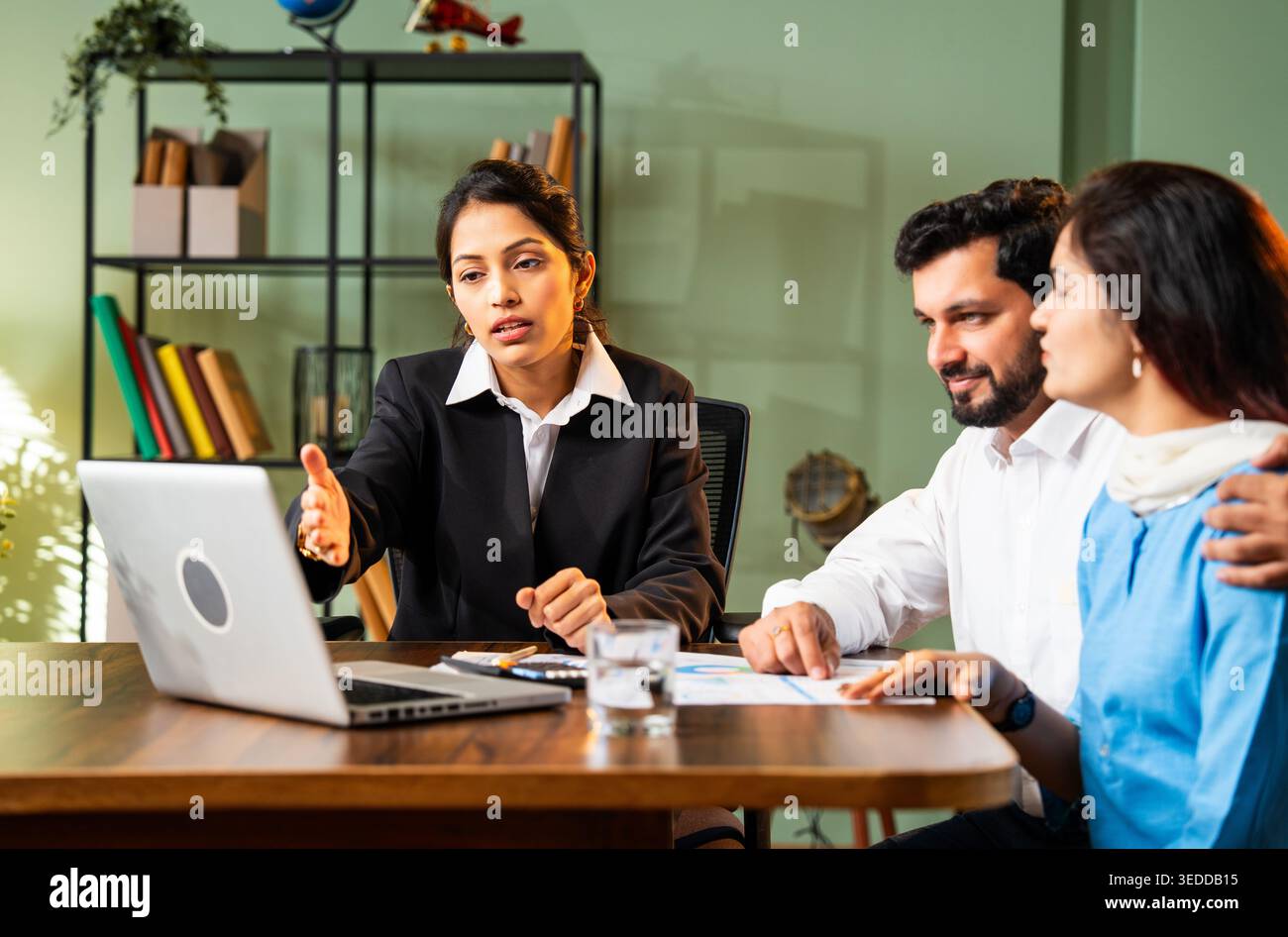 Cadre professionnel indien rencontrant un jeune couple dans un bureau moderne avec un ordinateur portable discutant des rapports financiers à travers la table et serrant la main sur R Banque D'Images