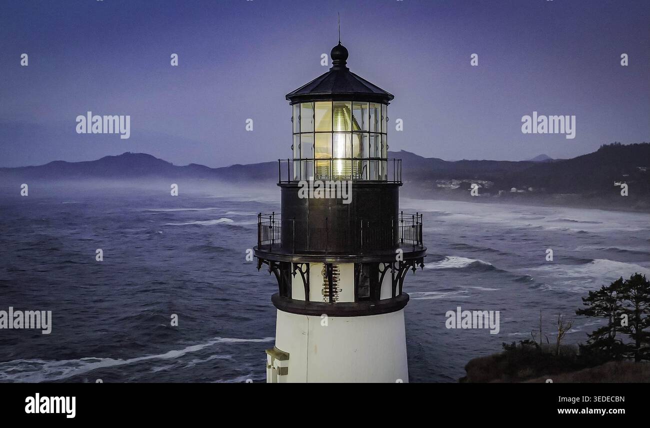 Vue aérienne du phare de Yaquina Head avec sa lanterne brillante au crépuscule surplombant les vagues écrasantes de l'océan Pacifique et les collines côtières brumeuses ne Banque D'Images
