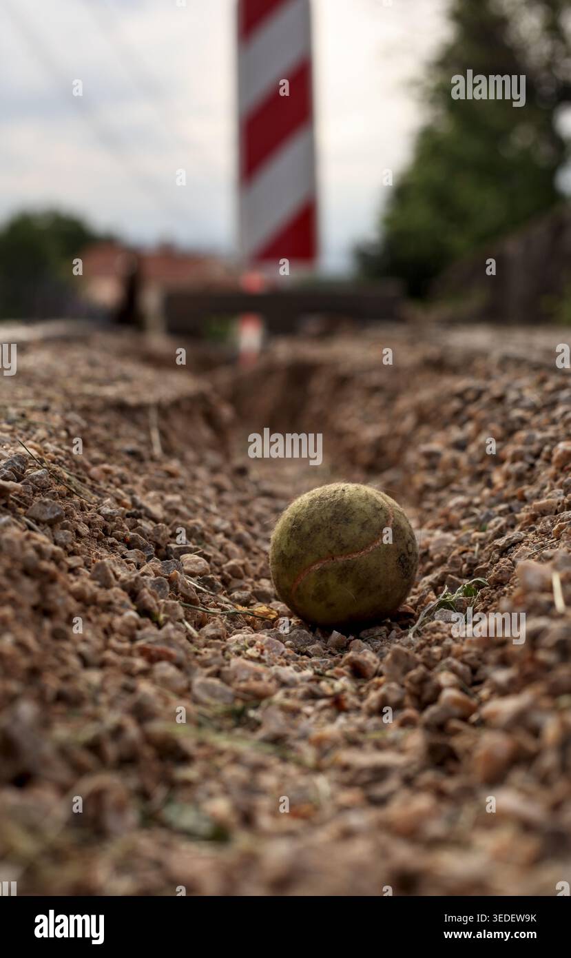 Vieille balle de tennis dans une rainure de terre sur un sol de gravier. Barrière routière rouge et blanche floue en arrière-plan sous un ciel couvert. Banque D'Images