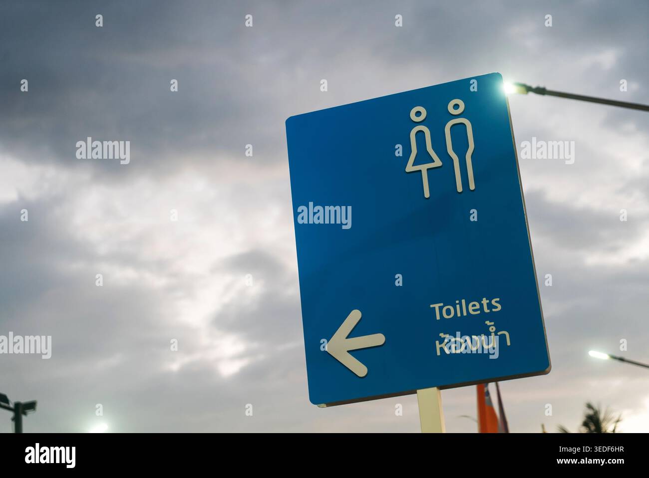 Un panneau directionnel bleu indiquant l'emplacement des toilettes publiques, avec des icônes universelles de genre et des instructions claires, placé contre un eveni nuageux Banque D'Images