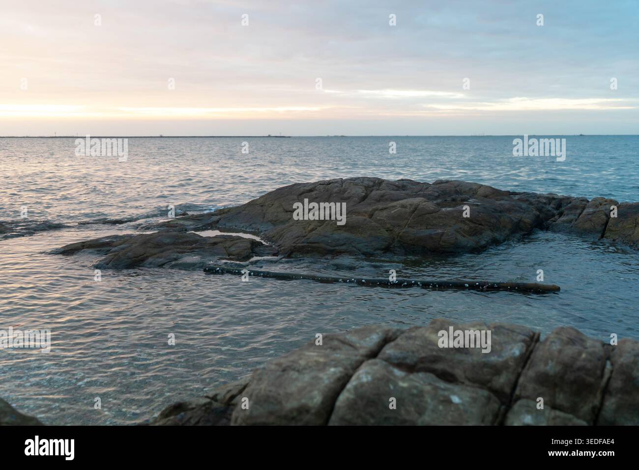 Une vue sereine sur l'océan au crépuscule, avec des rochers lisses dépassant des eaux calmes. Les teintes douces du ciel créent une atmosphère paisible, parfaite Banque D'Images