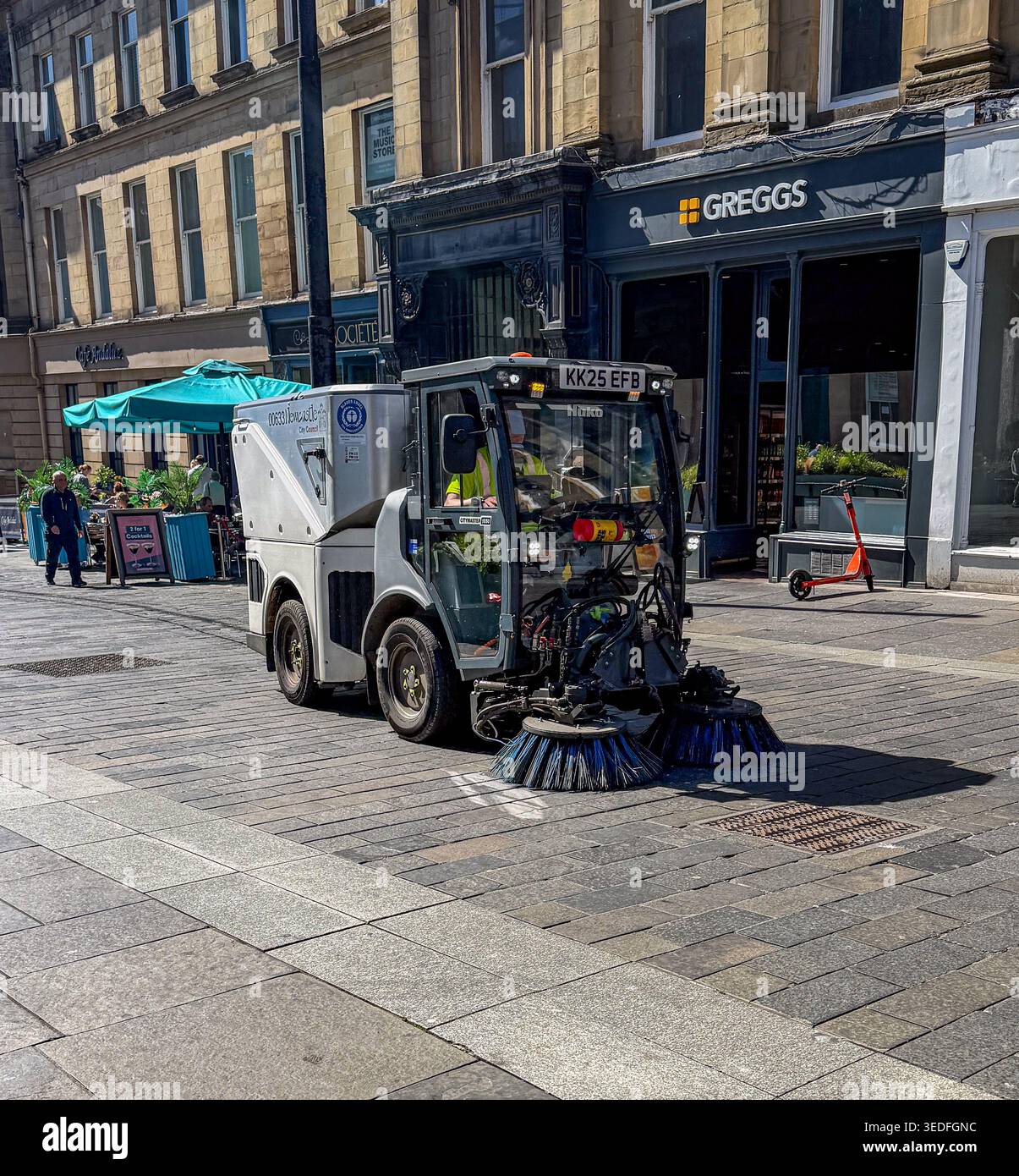 Un petit véhicule de balayage de route nettoyant une rue pavée dans un centre-ville avec des magasins et des sièges en plein air Newcastle upon Tyne - Image de stock capturée avec un smartphone