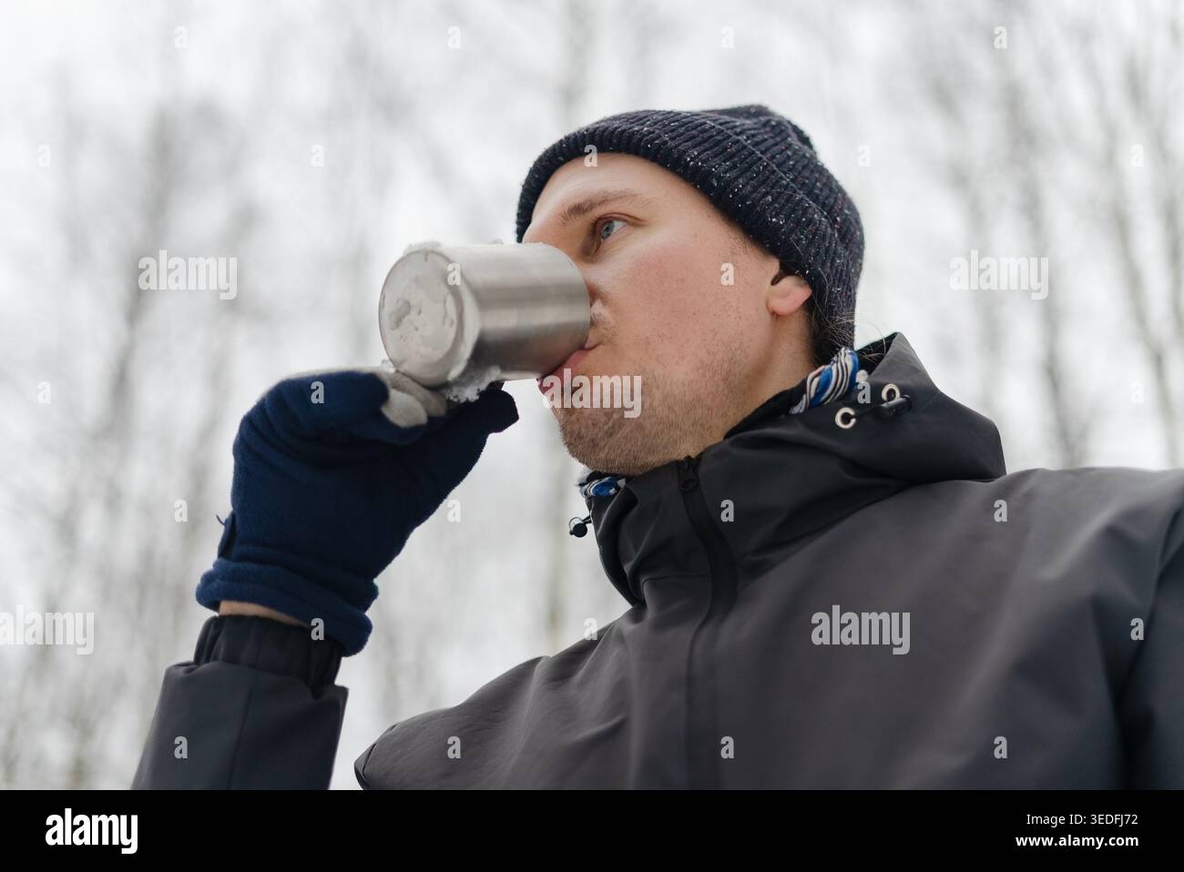 Homme buvant dans une tasse en métal portant une écharpe à motifs lors d'une froide journée d'hiver à l'extérieur. Un gars en bonnet sirotant une boisson chaude par temps neigeux. Confort chaleureux d'une boisson d'hiver sur une promenade glaciale Banque D'Images