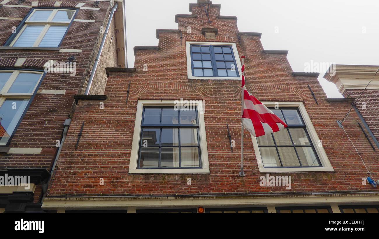 Bâtiment en brique avec un toit à pignon en escalier et un drapeau suspendu, présentant des détails architecturaux traditionnels le long d'une rue de la ville. Banque D'Images