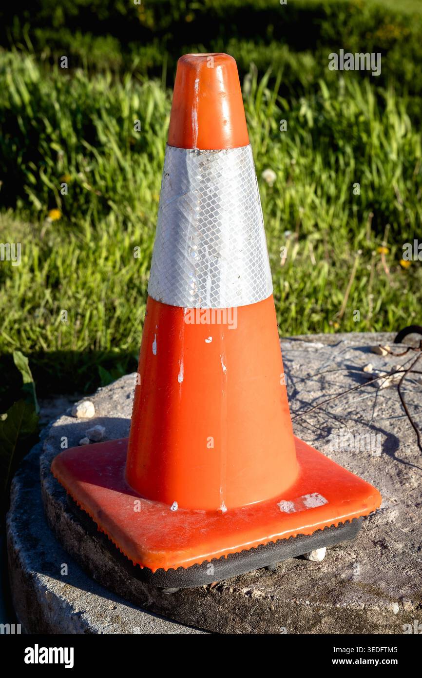 Cône de circulation orange avec bande réfléchissante assis sur le béton dans la lumière du soleil du soir avec fond d'herbe verte Banque D'Images
